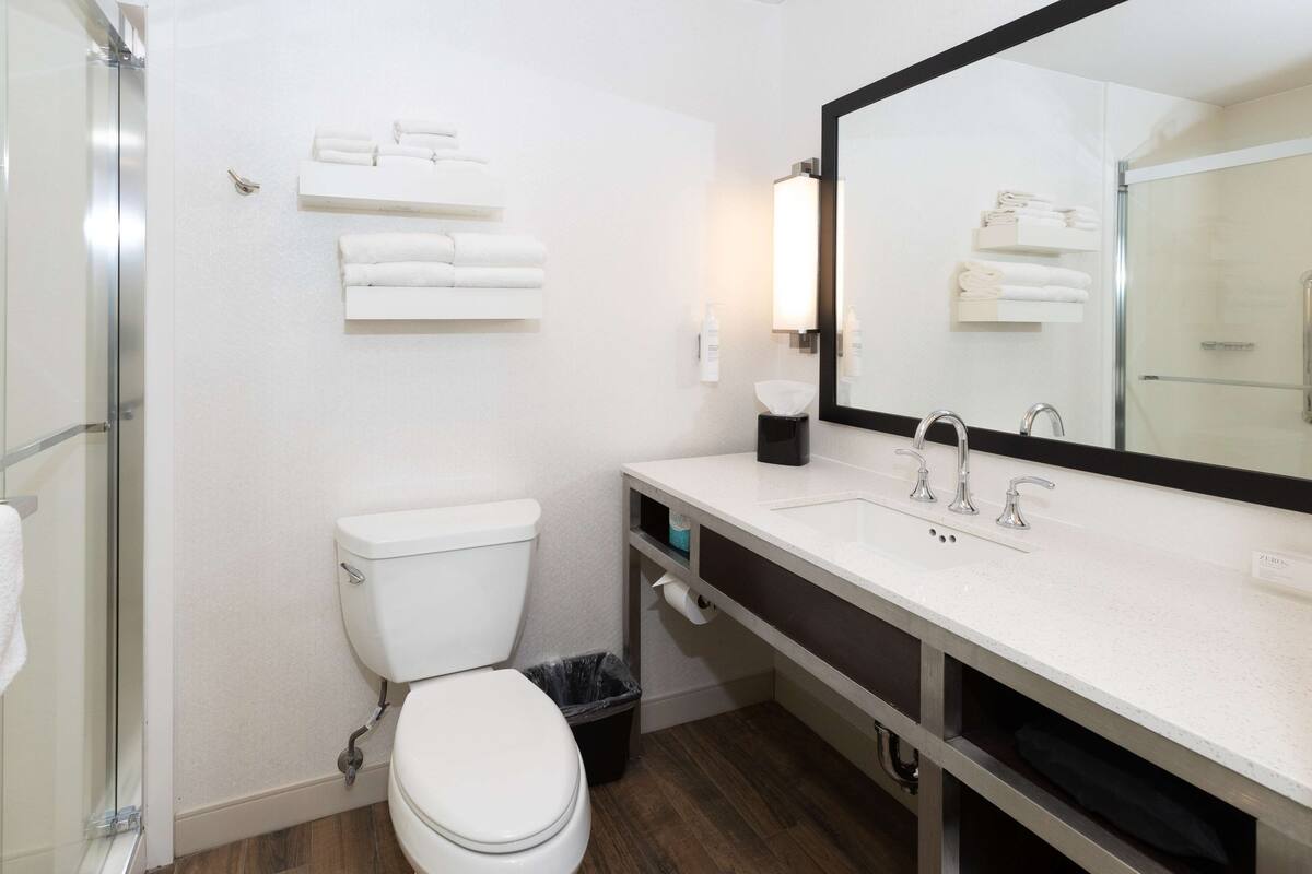 A bathroom with a toilet, sink, and shower. There are towels on a shelf and a trash can.