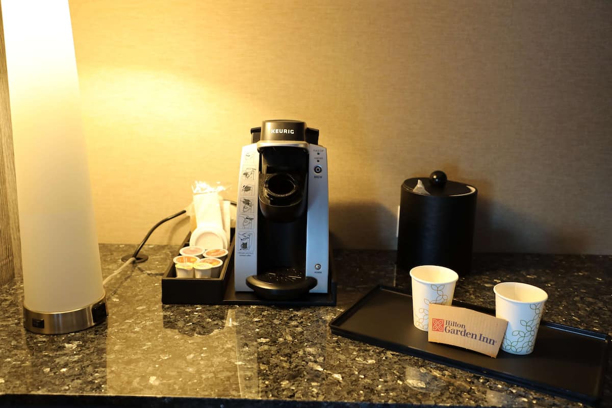 A Keurig coffee maker, a coffee dispenser, disposable cups, and a Hilton Garden Inn sign on a granite countertop.