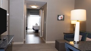 A hotel room with a bed, a desk with a lamp, a TV, and a view of the hallway. - Hilton Garden Inn Austin Airport (Austin)