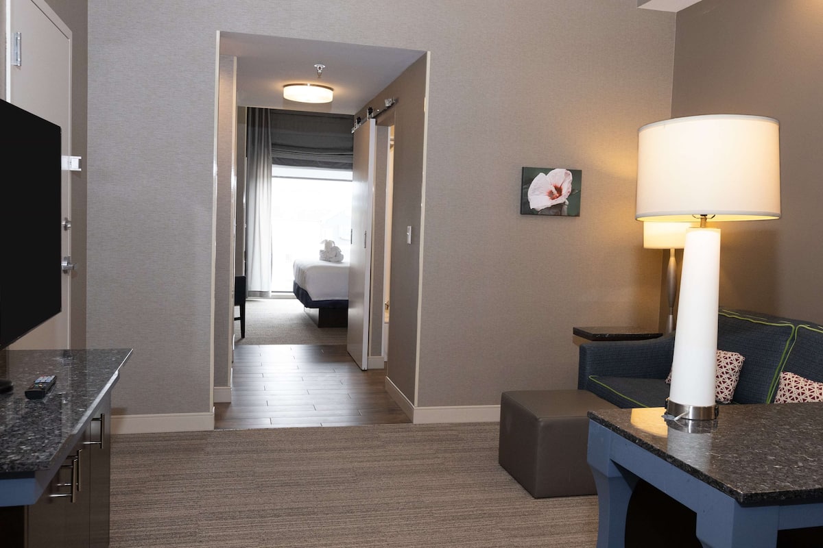 A hotel room with a bed, a desk with a lamp, a TV, and a view of the hallway.