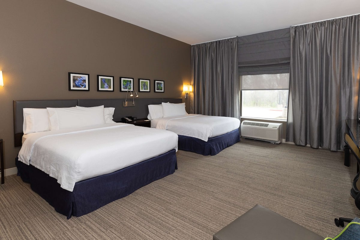 A hotel room with two beds, a desk, a chair, and a large window with curtains.