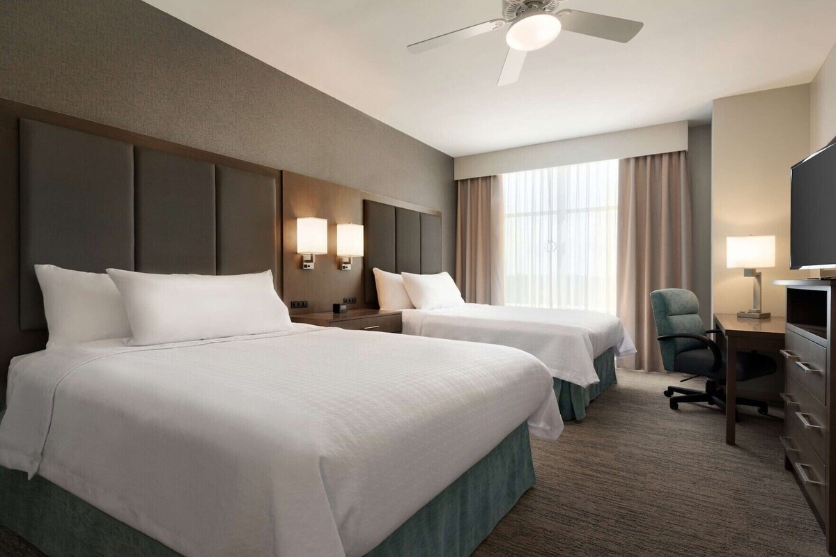 Een hotelkamer met twee bedden, een bureau, een stoel, een televisie en een plafondventilator.