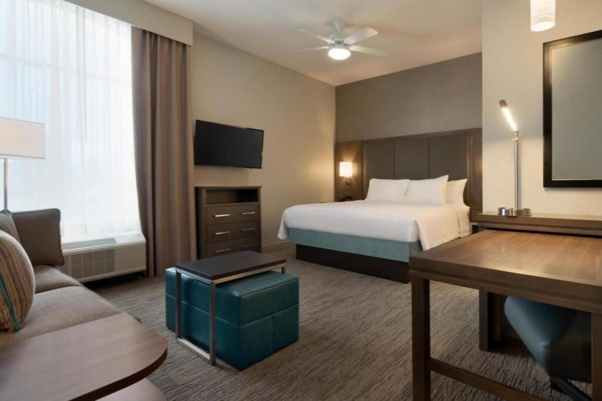 Une chambre d’hôtel équipée d’un lit, d’un bureau, d’une télévision, d’un ventilateur de plafond et d’une fenêtre avec des rideaux.