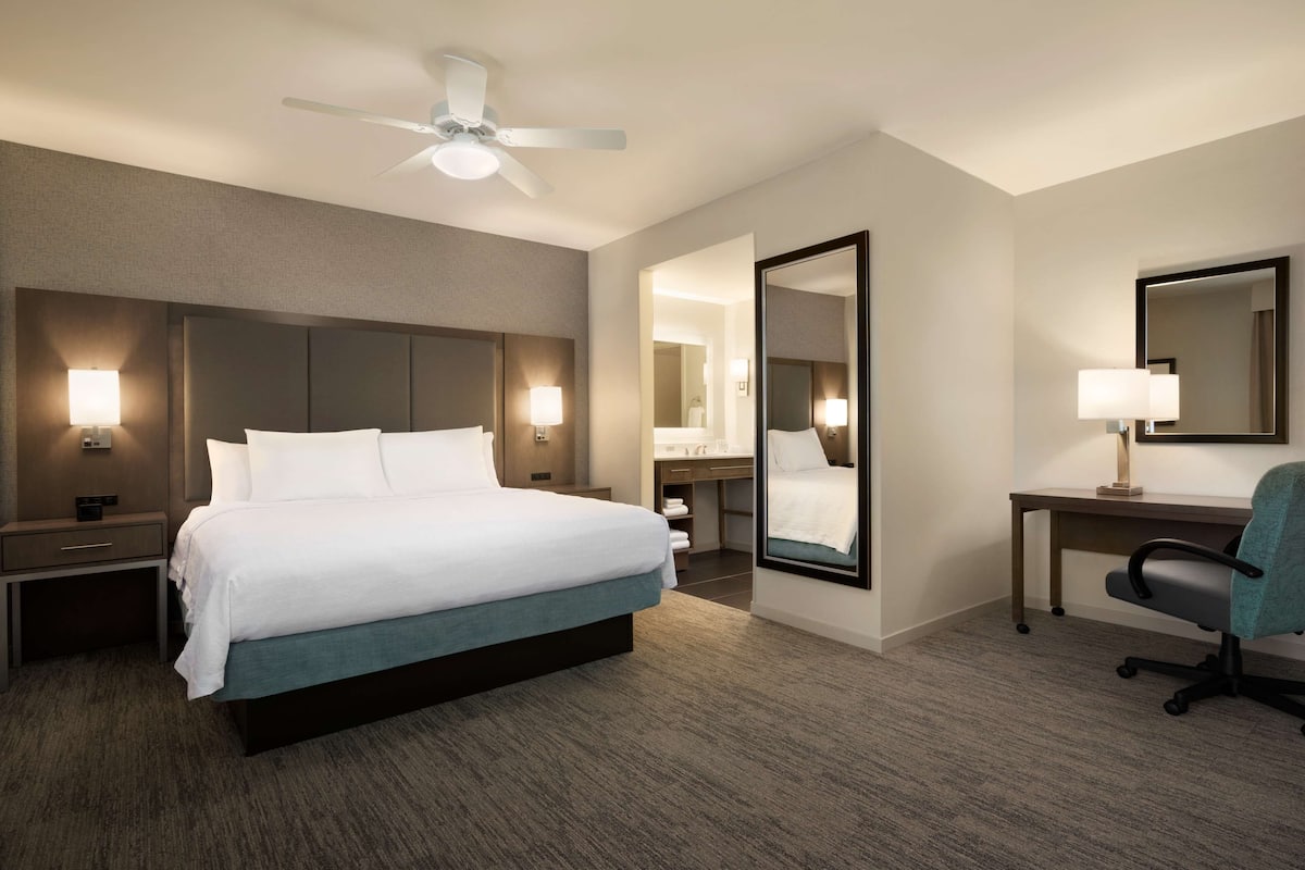 Une chambre d’hôtel avec un grand lit, des tables de chevet avec des lampes, un bureau avec une chaise, un ventilateur de plafond et un grand miroir.