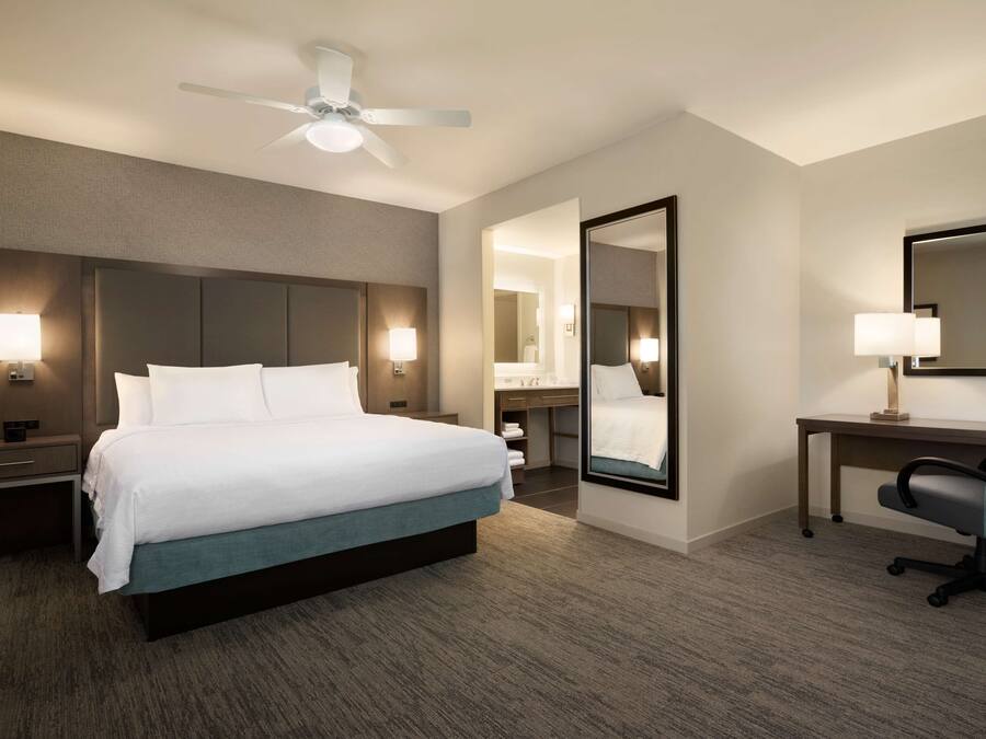 A hotel room with a large bed, bedside tables with lamps, a desk with a chair, a ceiling fan, and a large mirror.
