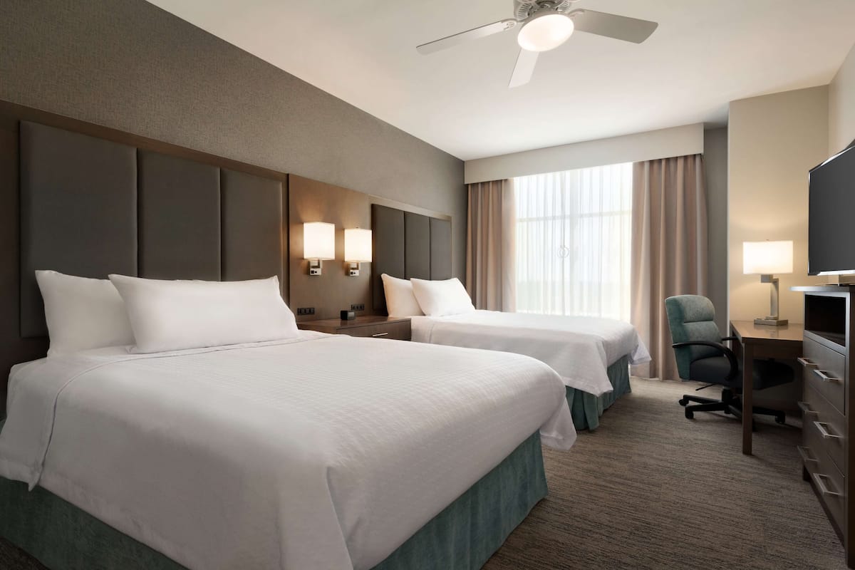 Une chambre d’hôtel avec deux lits, un bureau, une chaise, une télévision et un ventilateur de plafond.