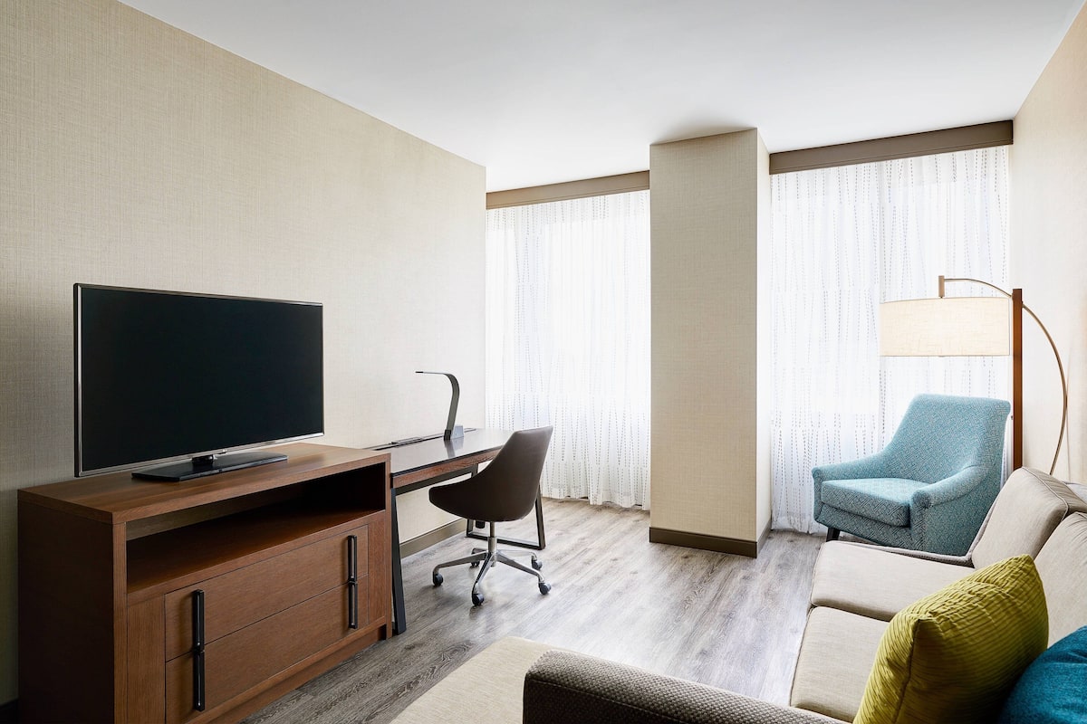 Une chambre d’hôtel avec un bureau en bois, une télévision, un fauteuil bleu, un canapé beige et une lampe sur pied.