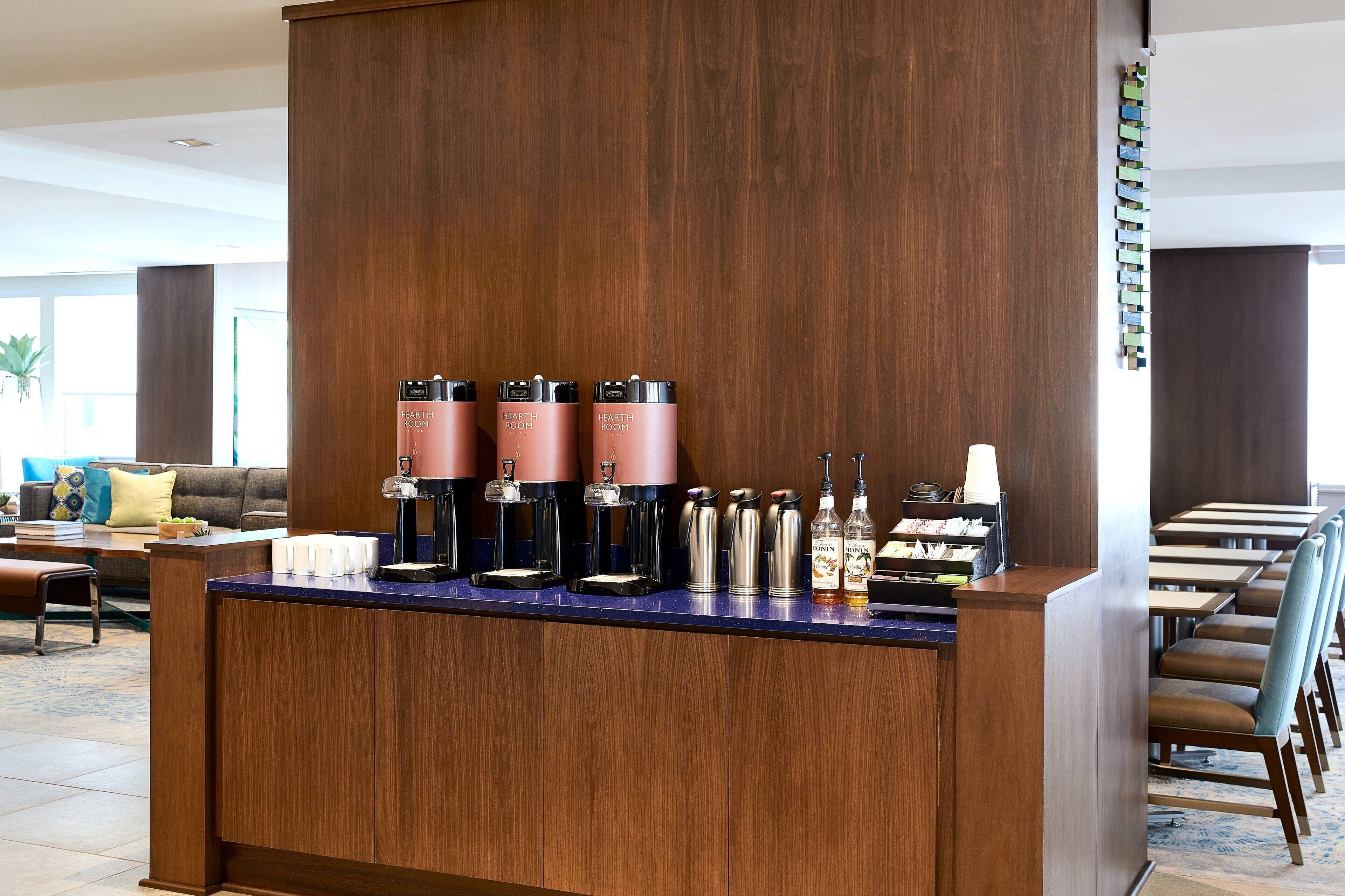 A hotel reception area with a wooden counter, multiple coffee machines, and a seating area with chairs and a sofa.