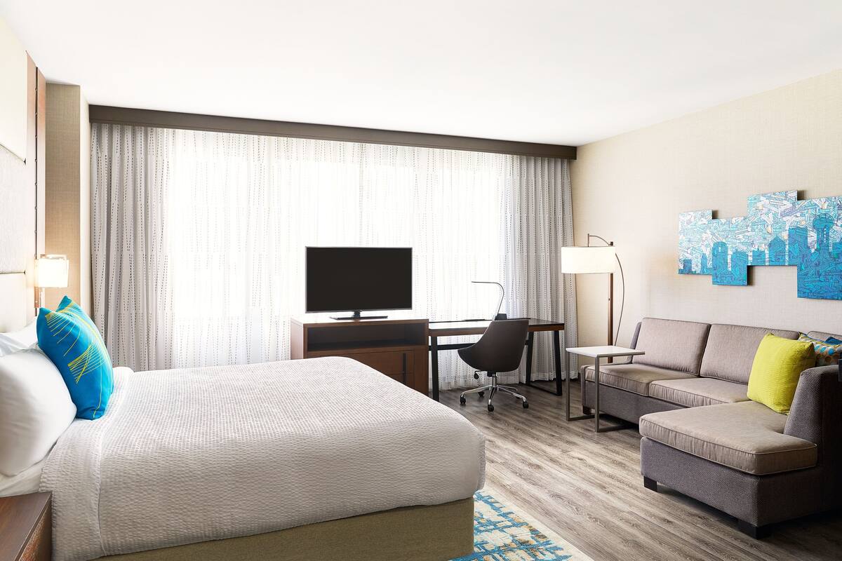Une chambre d’hôtel avec un lit, un bureau avec un ordinateur, un canapé et un tableau représentant un paysage urbain accroché au mur.