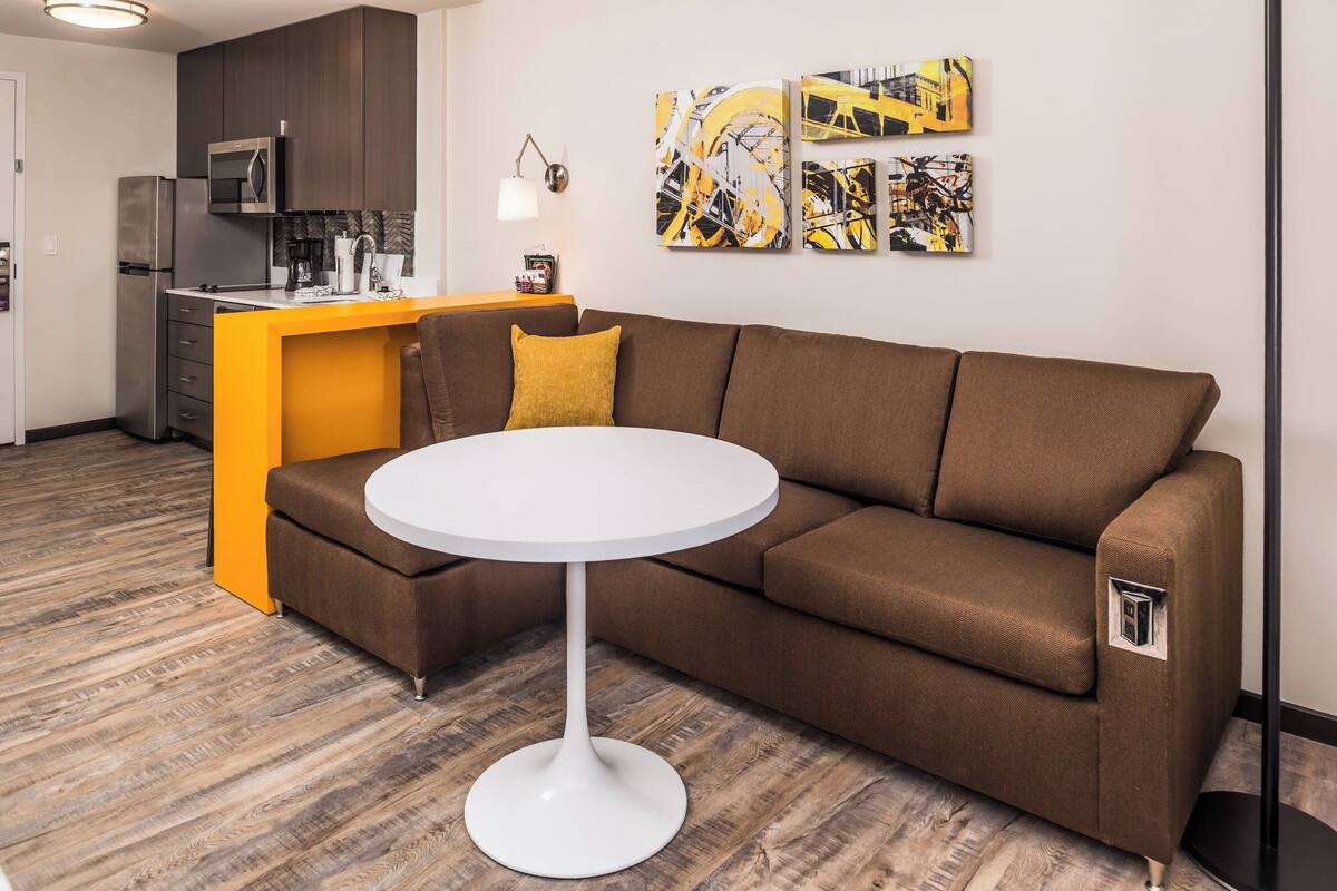 A modern living space with a brown sectional sofa, a white round table, and a kitchen area with a microwave and coffee maker.