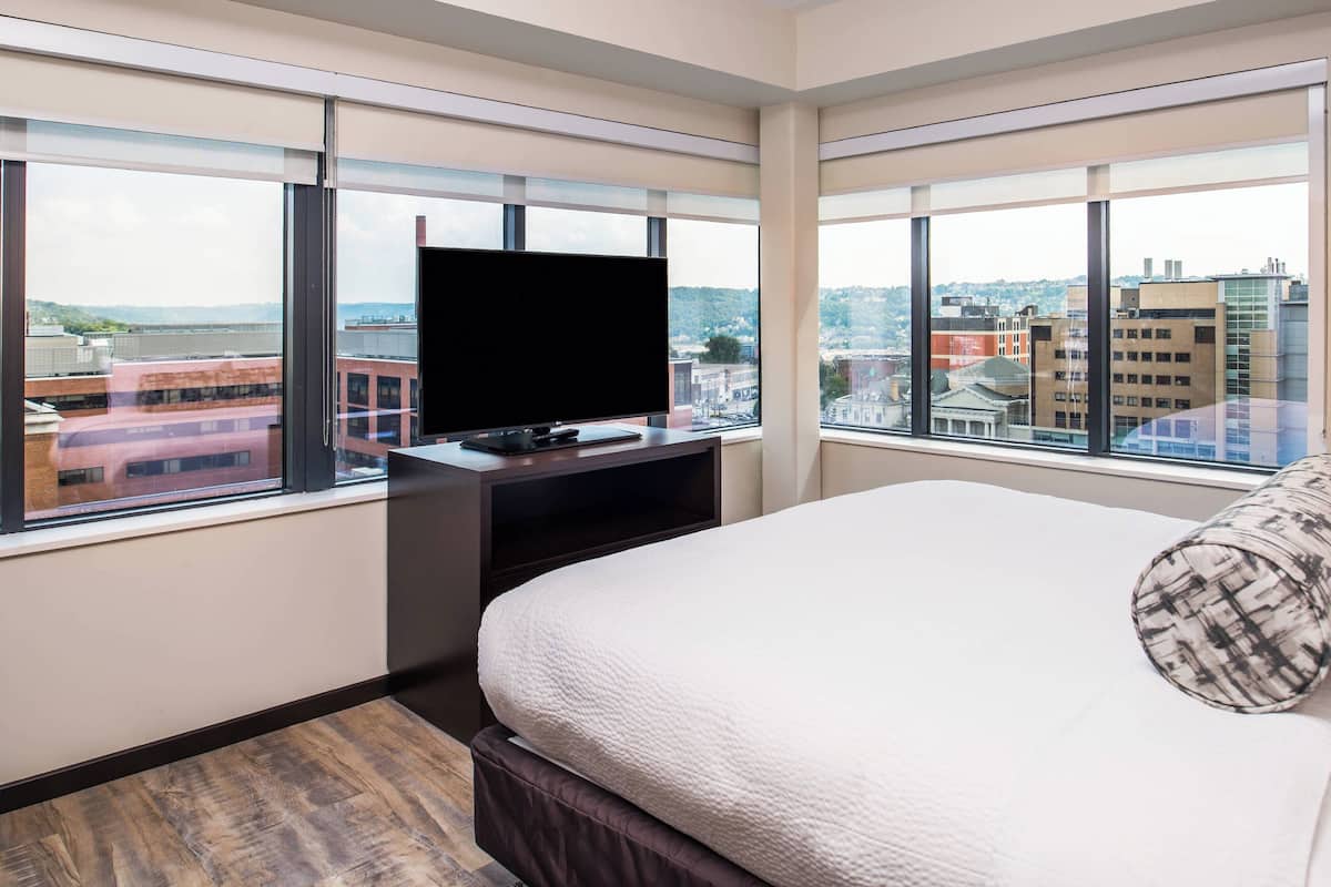 A hotel room with a large bed, a flat-screen TV on a stand, and a city view through large windows.