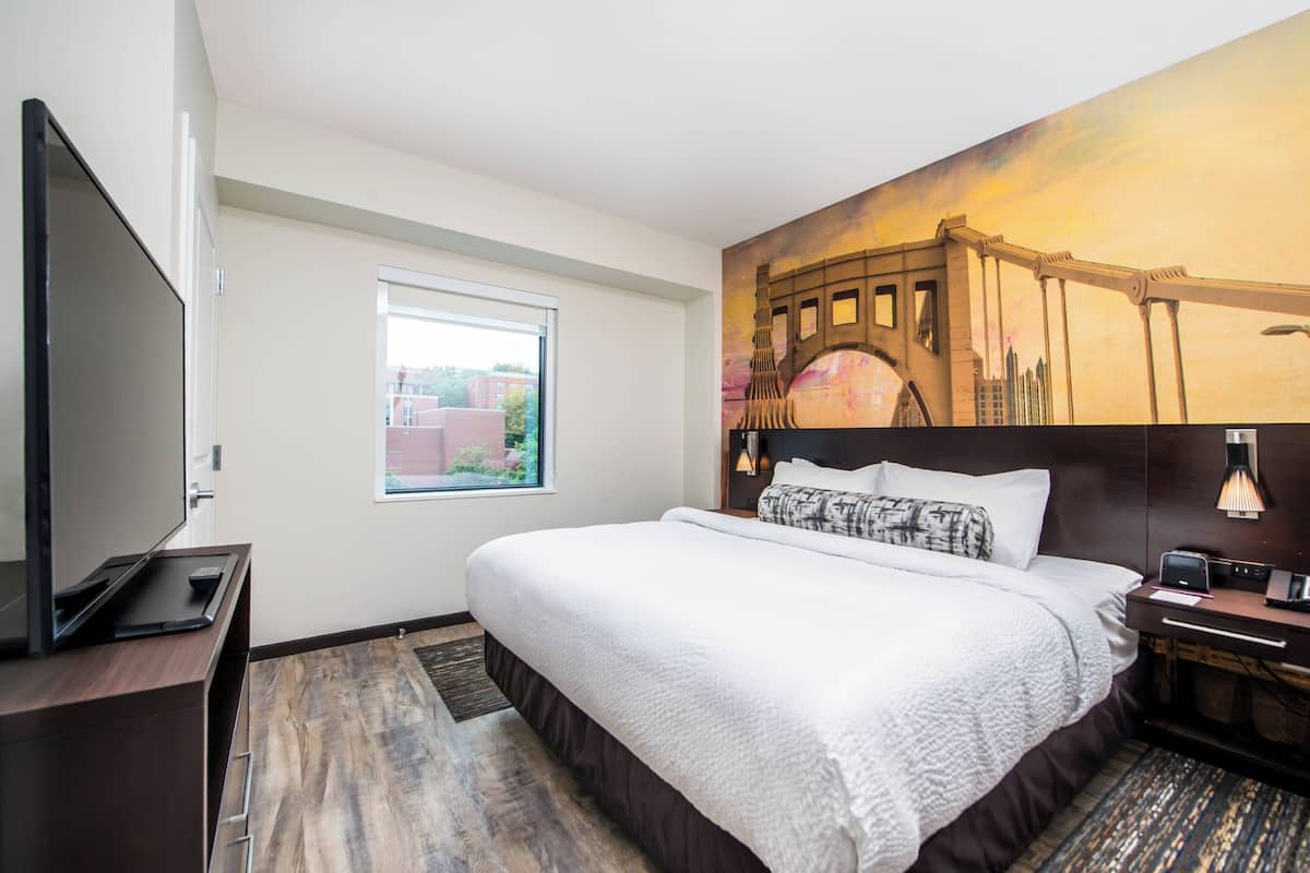 A hotel room with a large bed, a flat-screen TV, a window with a view of buildings, and a mural of a bridge.