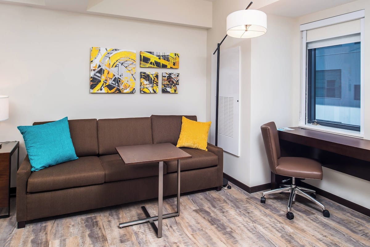 A modern living room with a brown sofa, a small table, a desk with a chair, and abstract art on the wall.
