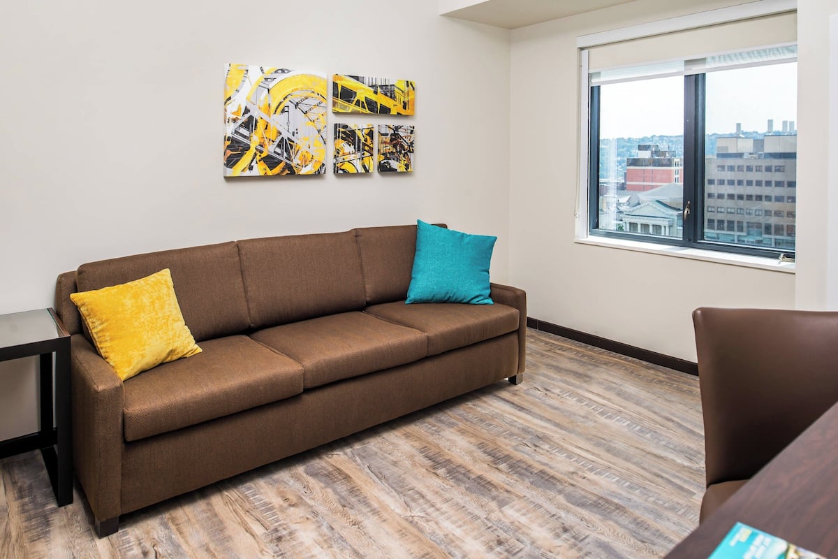 A modern living room with a brown sofa, two cushions, a small table, and a large window with a city view.