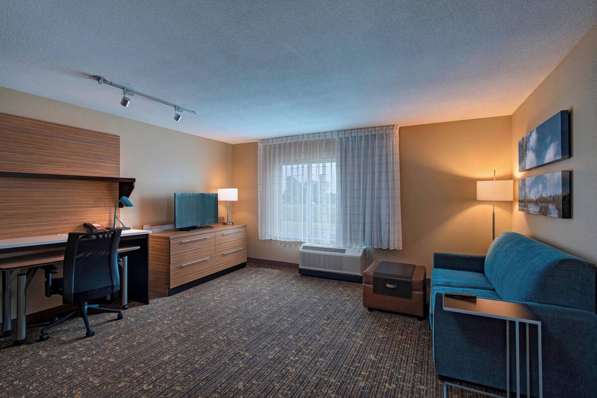 Ein Hotelzimmer mit Schreibtisch, einem blauen Sofa, einer Lampe, einem Fenster mit Vorhängen und einem Bild an der Wand.
