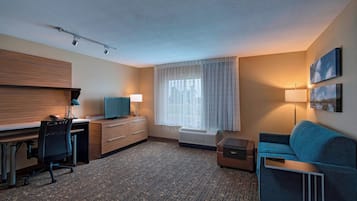 Habitación de hotel con escritorio, sofá azul, lámpara, ventana con cortinas y un cuadro en la pared.