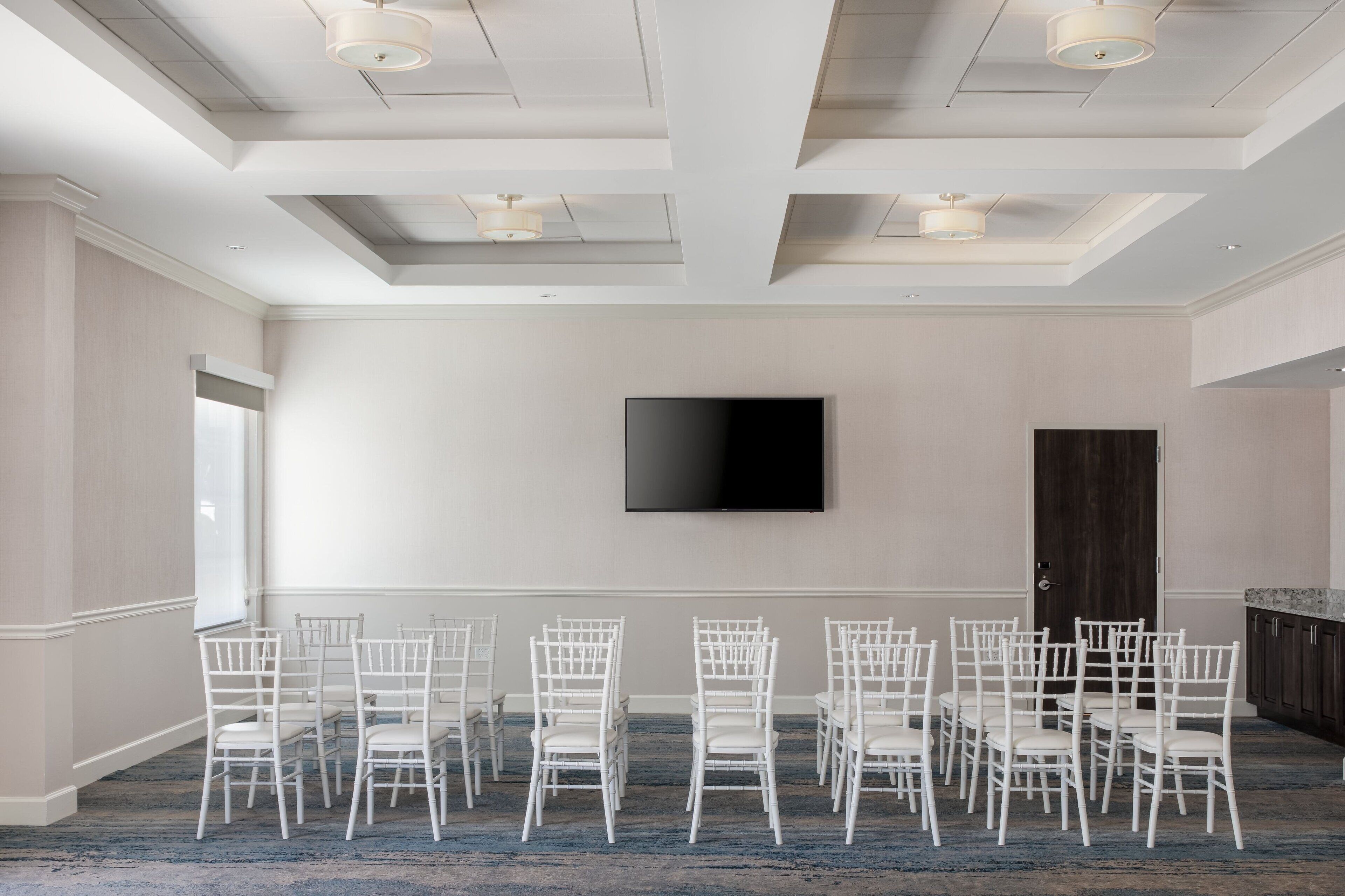 Un salón de conferencias con sillas blancas dispuestas en filas, un televisor de pantalla plana en la pared y una puerta de madera oscura.