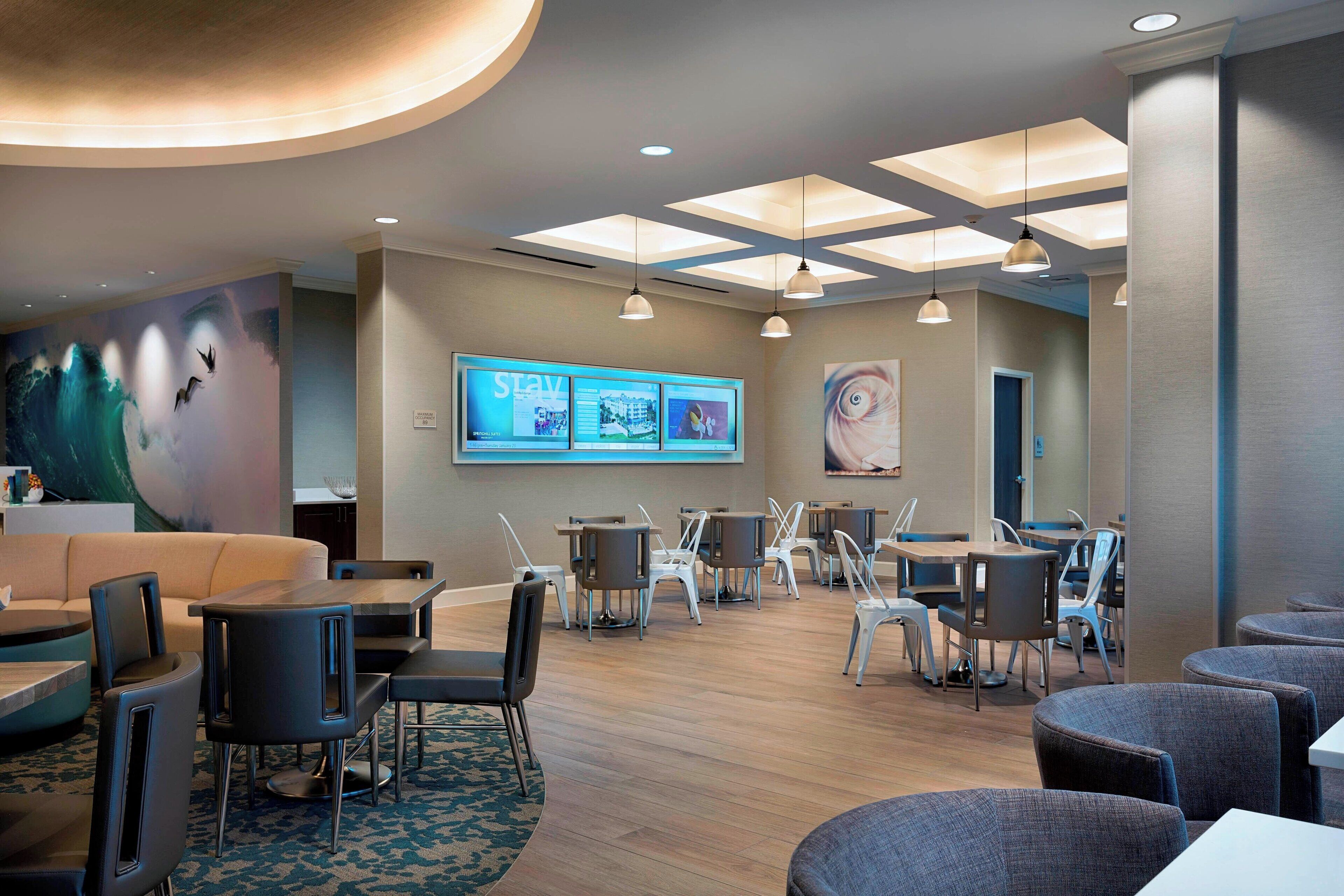 A modern hotel lobby with a central seating area, a large digital display, and a ceiling with skylights.