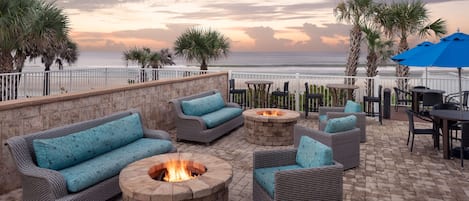 A patio with wicker furniture, a fire pit, and a view of the ocean at sunset.