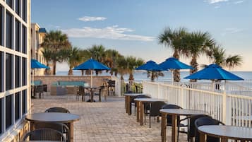 An outdoor patio with tables and chairs, blue umbrellas, and palm trees.