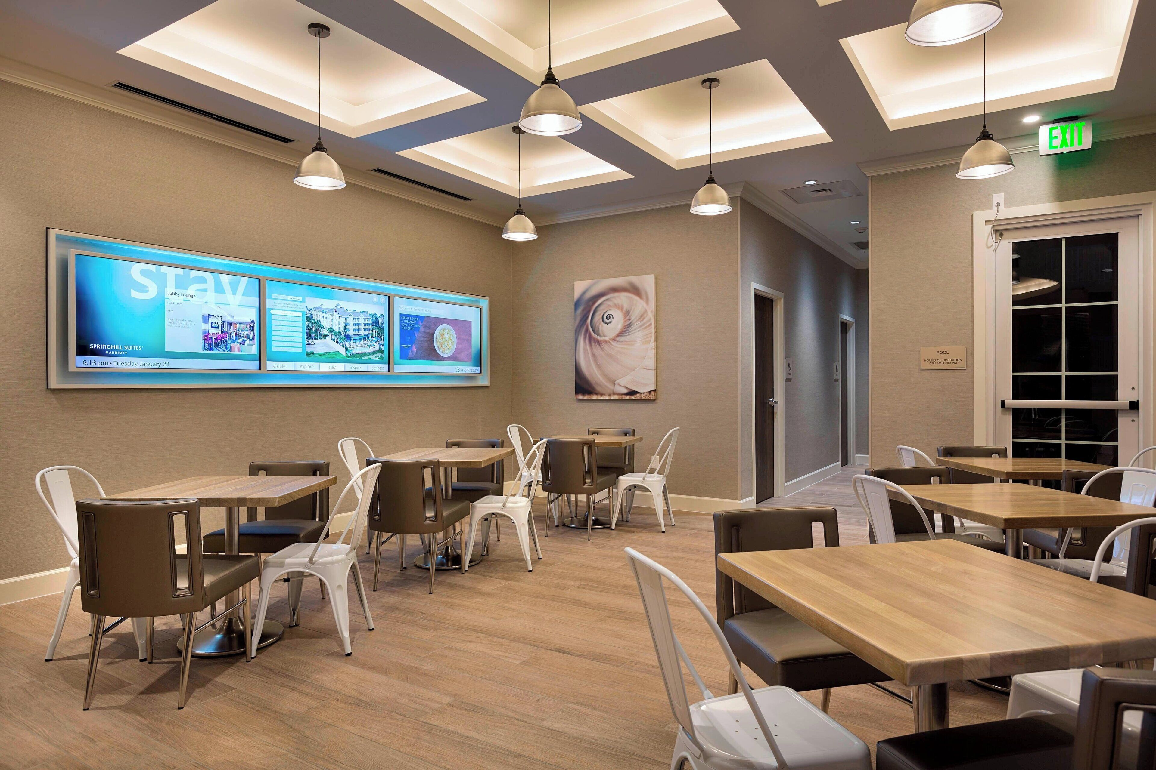 A conference room with multiple tables and chairs, a large screen displaying information, and an exit sign.