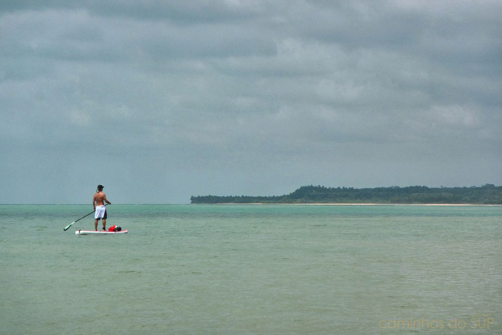 Una persona haciendo paddleboard en un mar tranquilo, con una isla y una playa a lo lejos.
