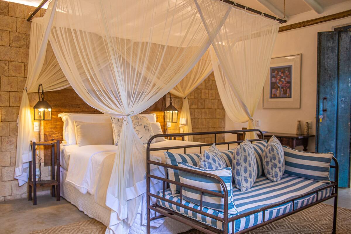 A four-poster canopy bed with white linens and striped pillows, a stone wall, and two wall-mounted lamps.