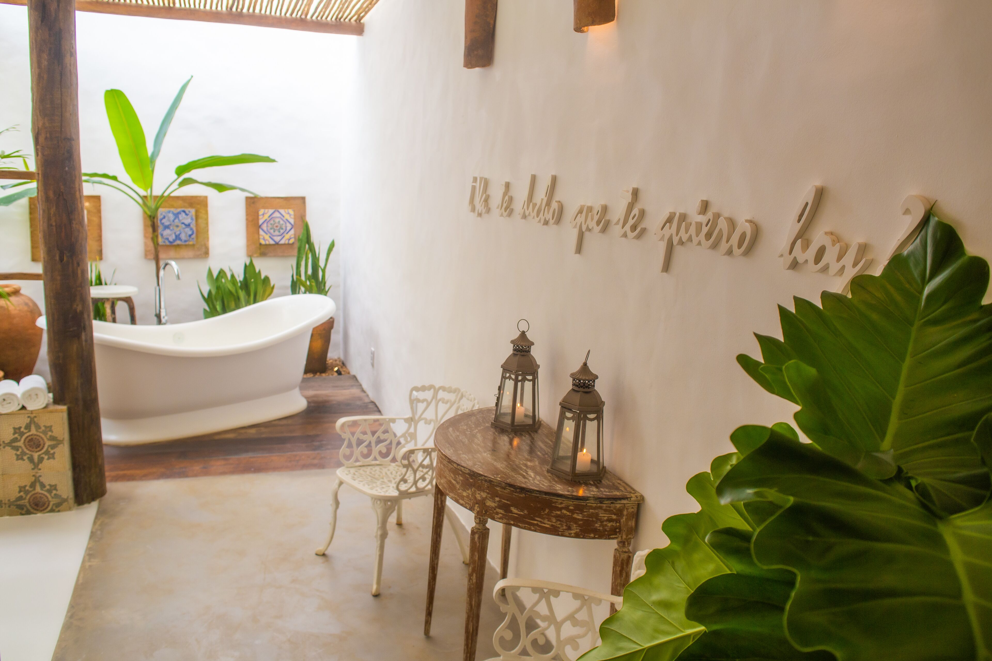 Un baño con una bañera blanca, sillas blancas, una mesa de madera con faroles y una pared decorada con una inscripción.