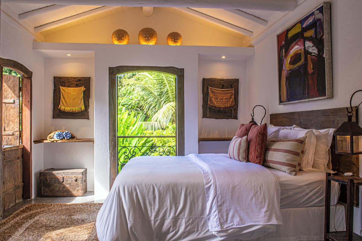 A bedroom with a large bed, a window with a view of greenery, a wooden chest, and a painting on the wall.