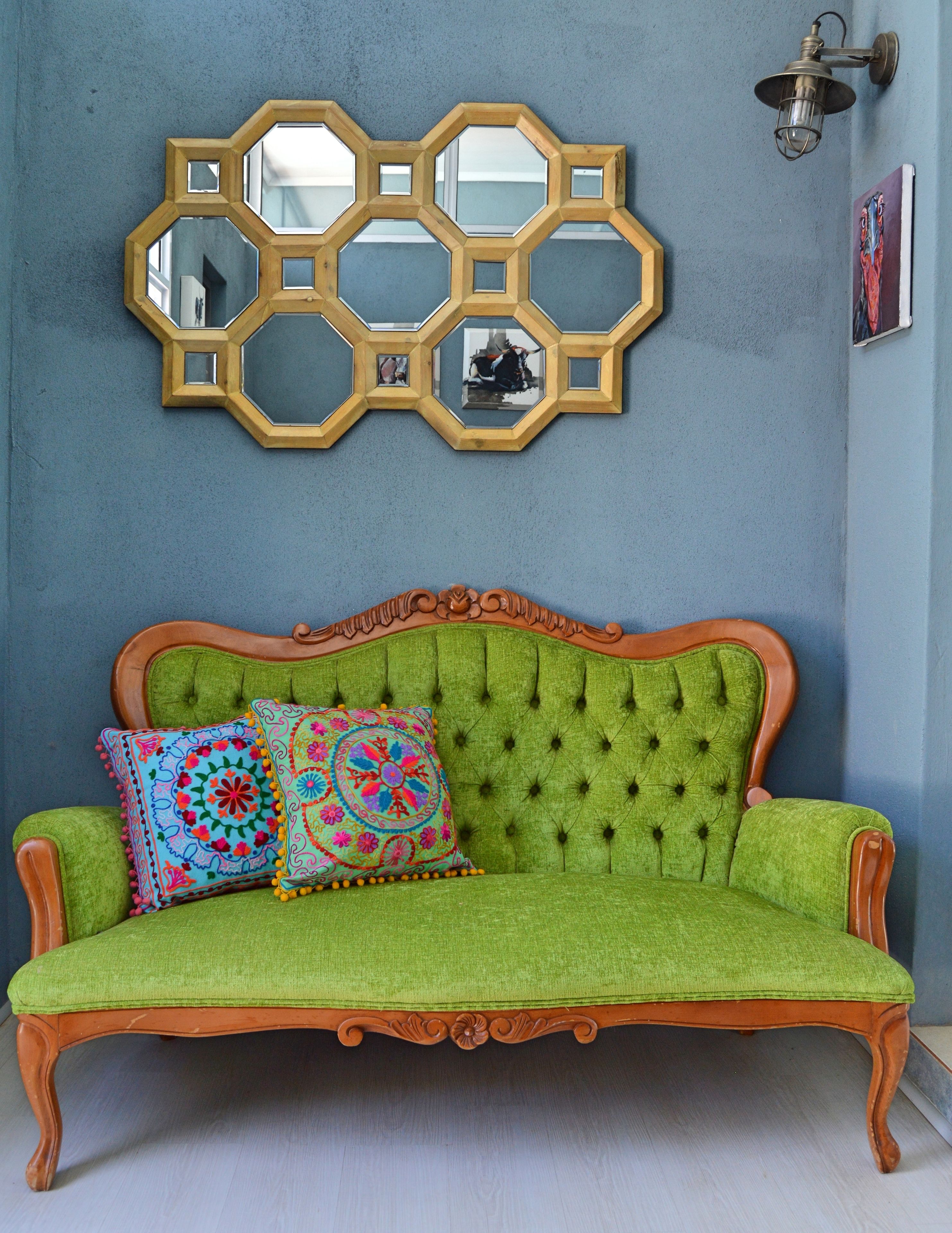 A green upholstered sofa with ornate wooden carvings, two colorful patterned cushions, and a hexagonal mirror above it.