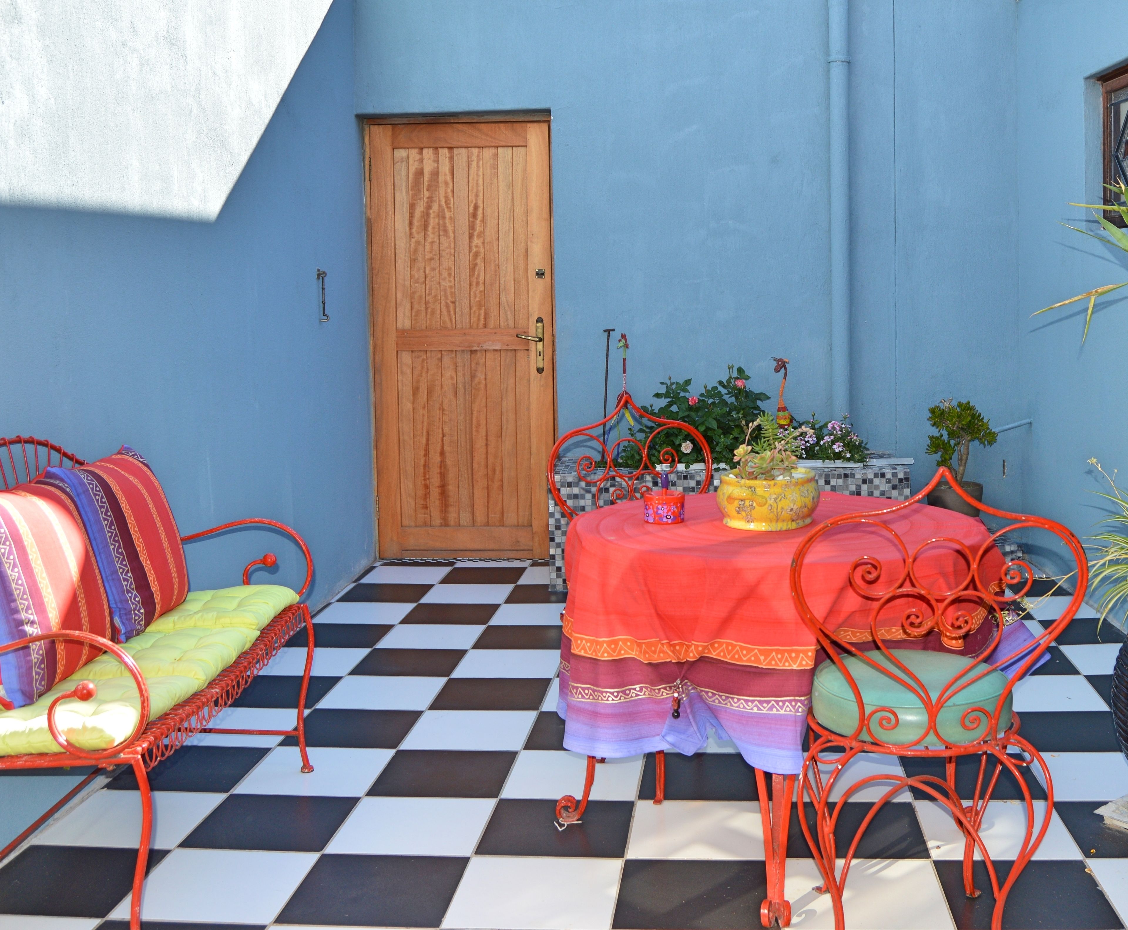 A room with a checkered floor, a wooden door, a table with a red tablecloth, and metal chairs with colorful cushions.