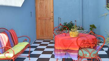 A room with a checkered floor, a wooden door, a table with a red tablecloth, and metal chairs with colorful cushions.