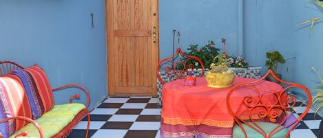 A room with a checkered floor, a wooden door, a table with a red tablecloth, and metal chairs with colorful cushions.