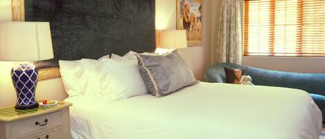 A neatly made bed with white and grey pillows, a bedside table with a lamp, and a chair with a teddy bear.
