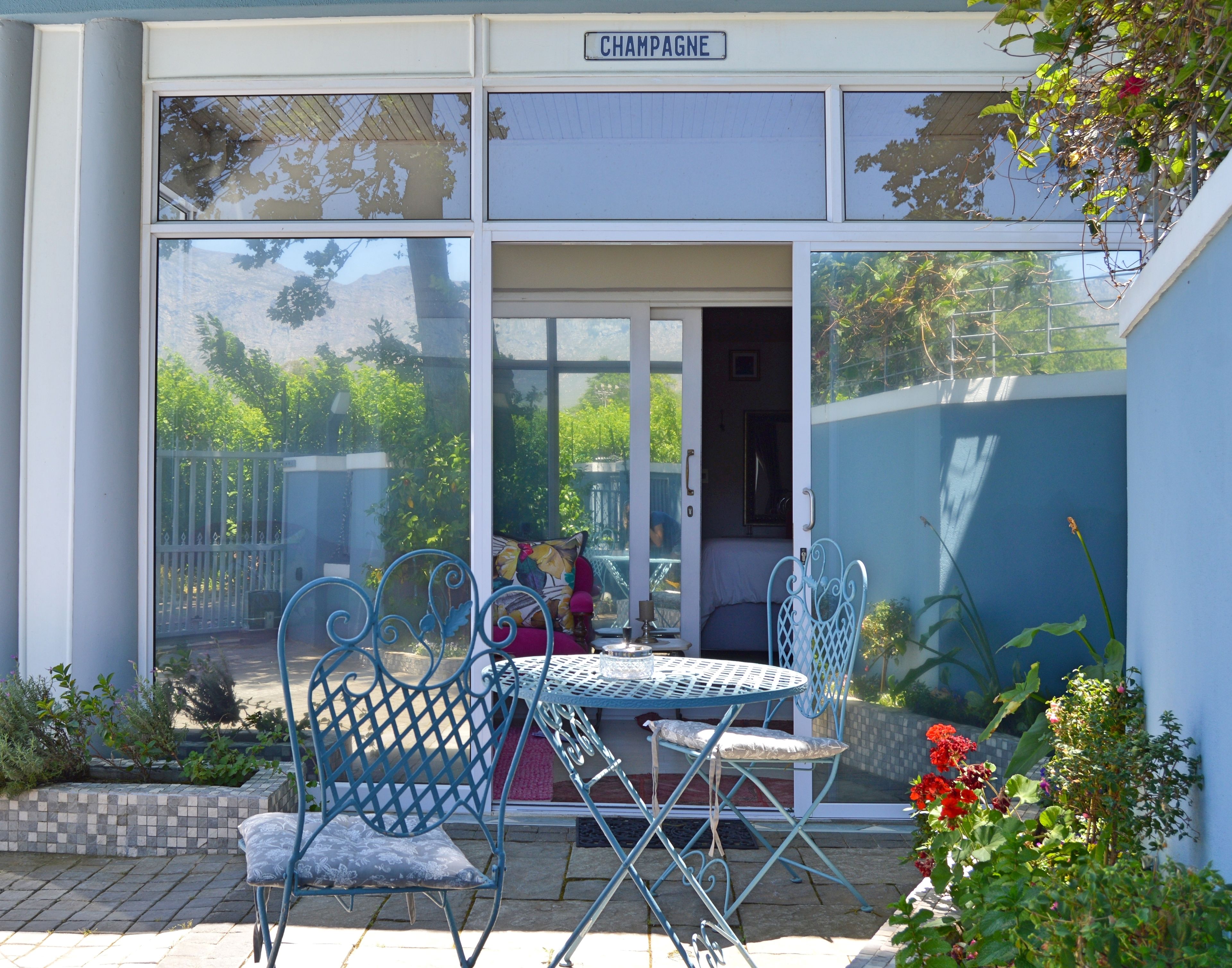 A covered outdoor seating area with metal chairs and a table, a door leading inside, and a view of greenery through the glass panels.