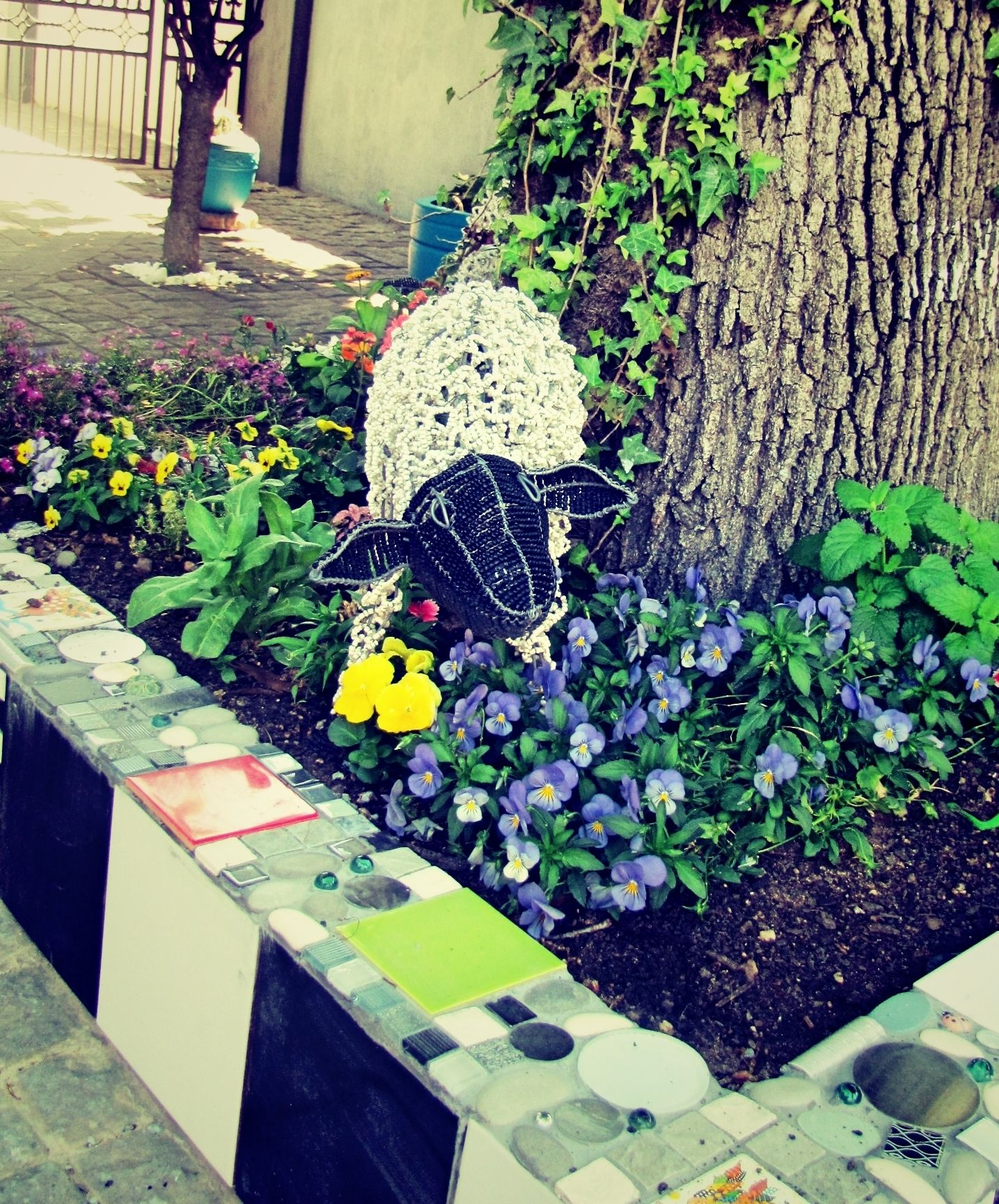 Una jardinera con una escultura de crochet, diversas flores y un árbol.