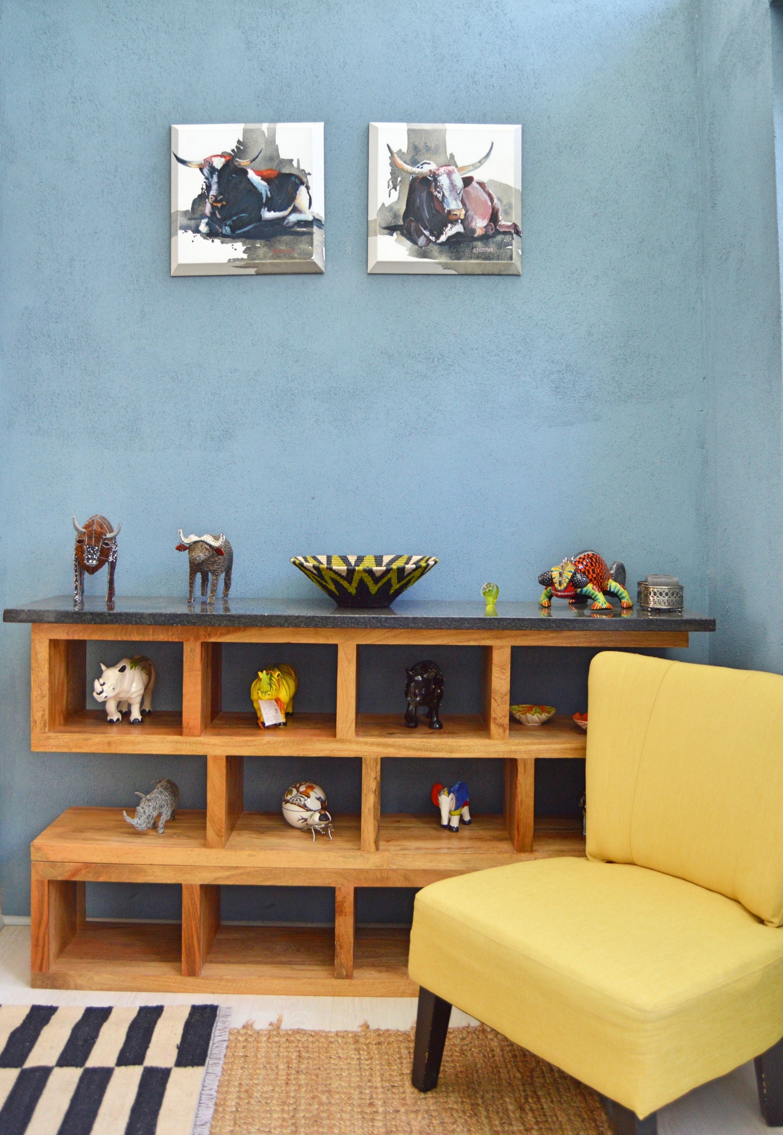 A room with a blue wall, two paintings of bulls, a wooden shelf with various animal figurines, a yellow chair, and a striped rug.