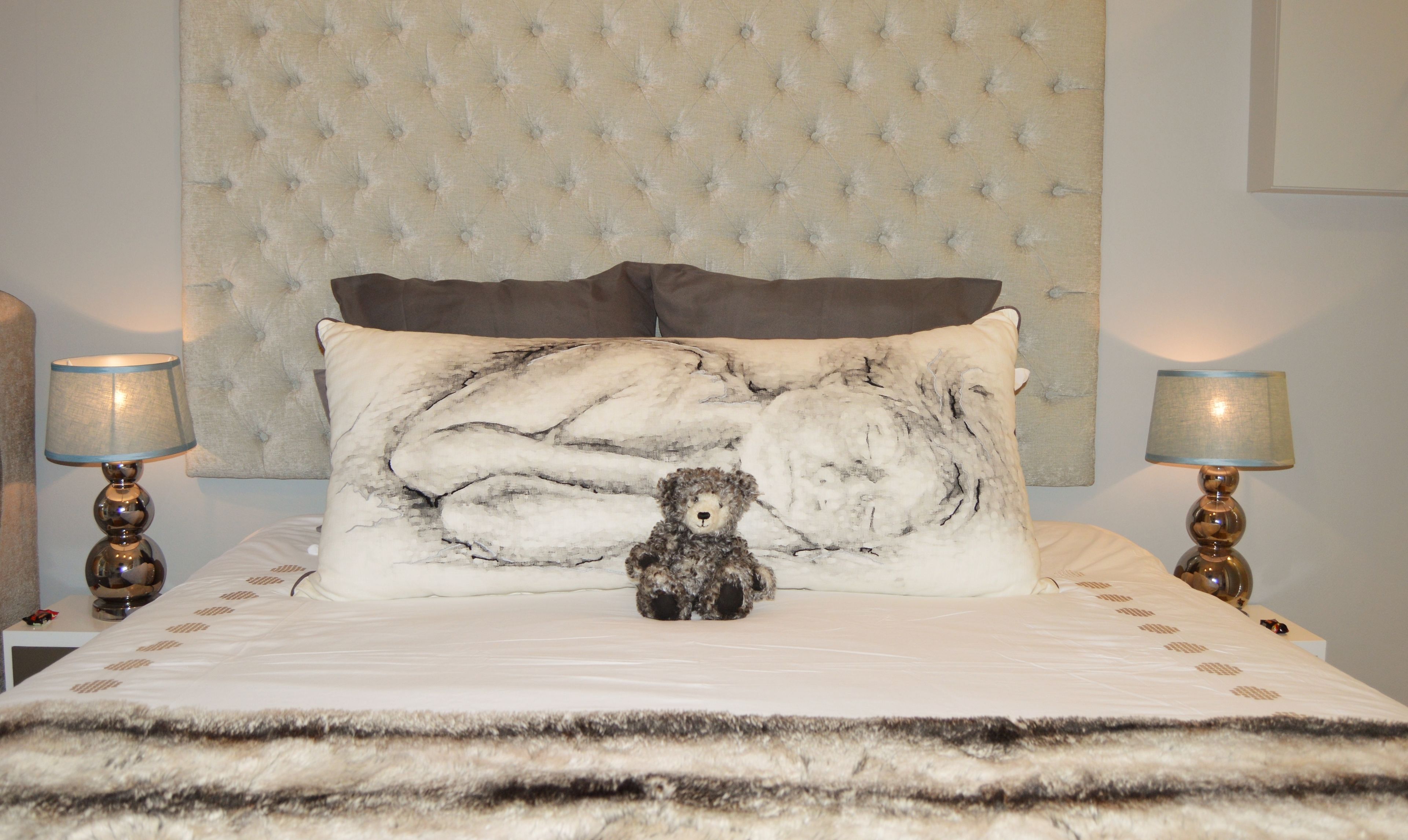 Una cama con cabecera capitonada, dos lámparas de noche y un cojín decorativo con un diseño de oso.