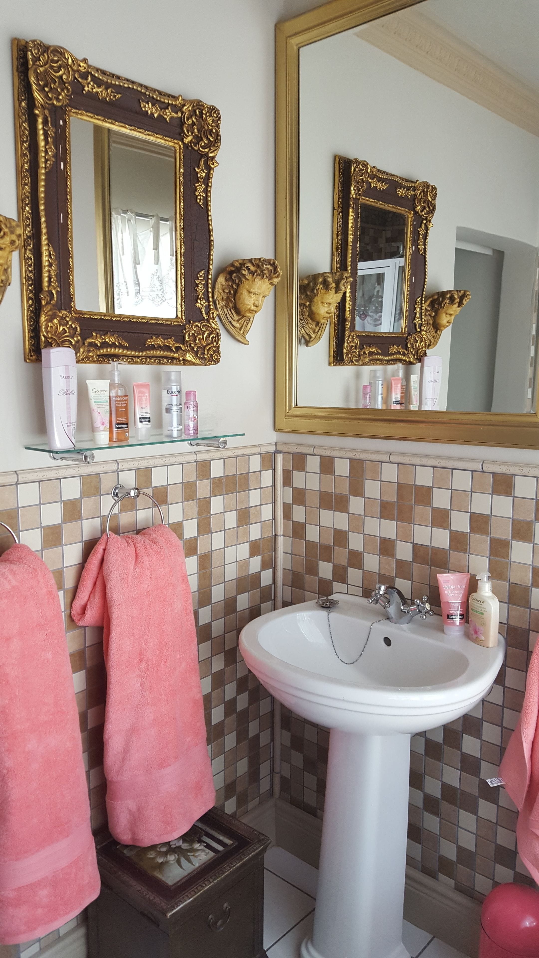 Baño con lavamanos blanco, dos espejos con marcos dorados ornamentados, repisas con artículos de aseo y toallas rosas.