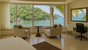 A hotel room with two armchairs, a small table, a desk, and a large window offering a view of a mountainous landscape and a body of water. - Villa Armonia Luxury Suites (Puerto Vallarta)