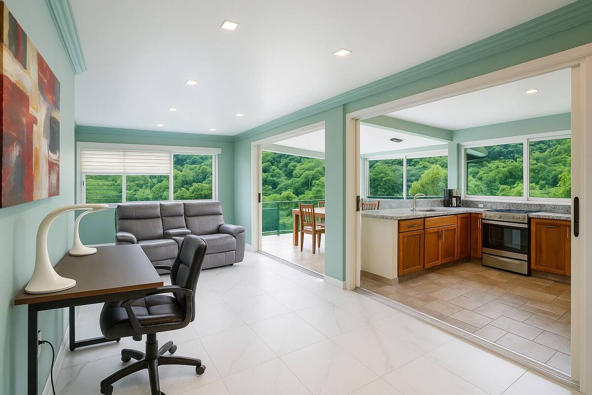 A spacious living area with a sofa, a desk with a chair, and a dining table. There is a kitchen with a stove and a view of greenery outside.