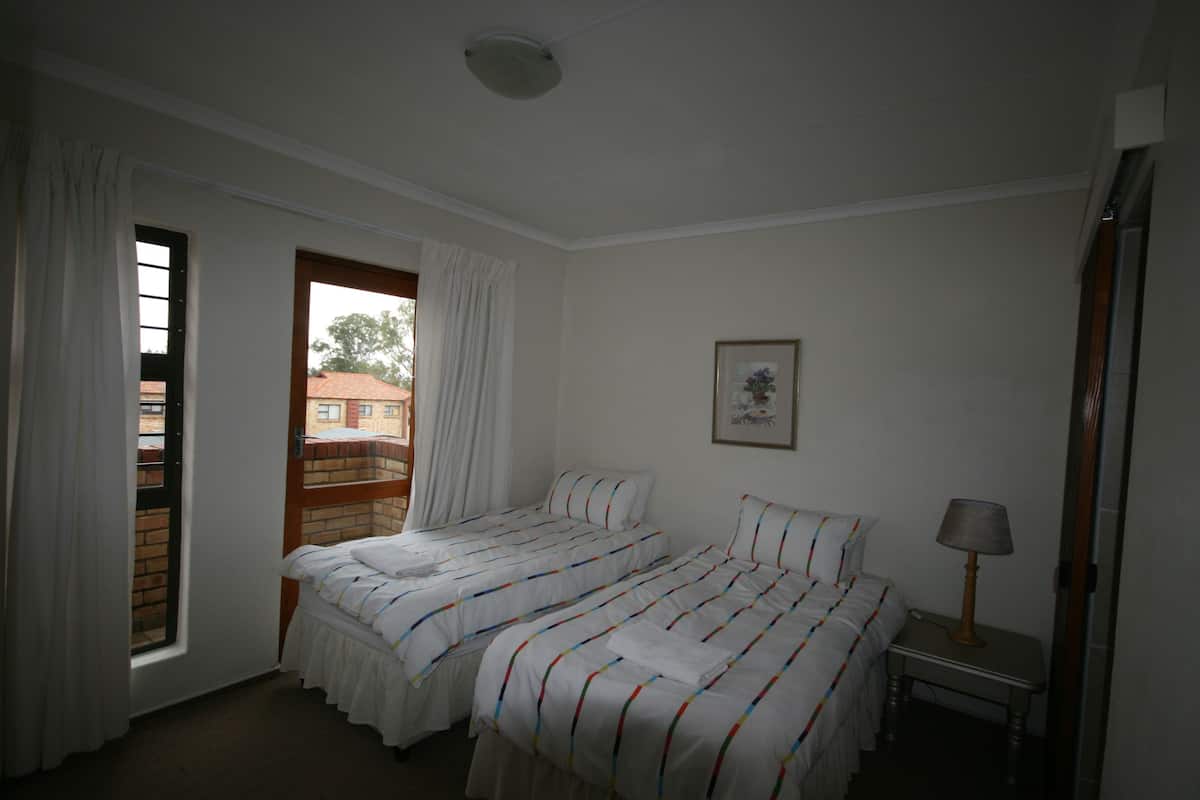 A room with two beds, a window with curtains, a framed picture on the wall, and a bedside table with a lamp.