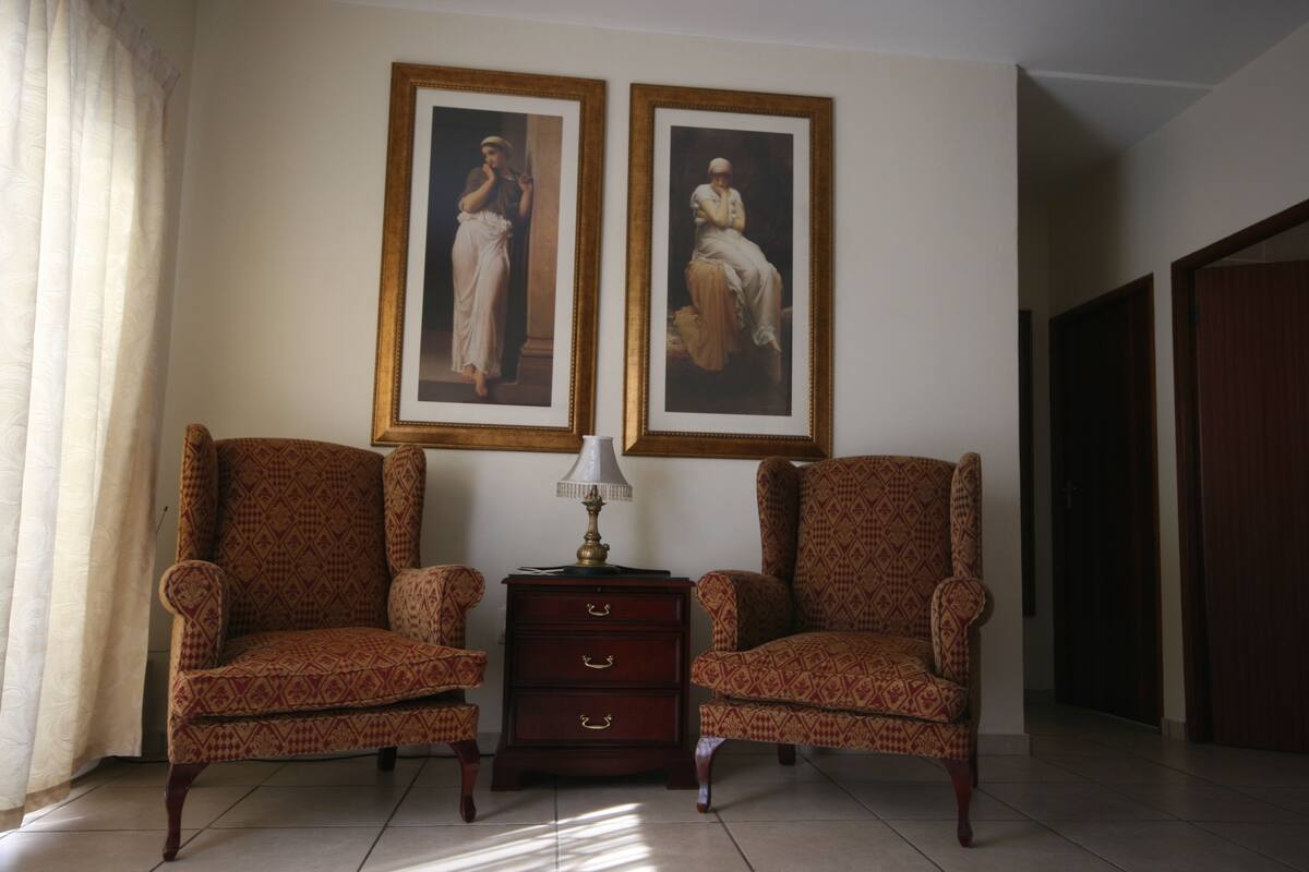 A room with two patterned armchairs, a small wooden side table with a lamp, and two framed paintings on the wall.
