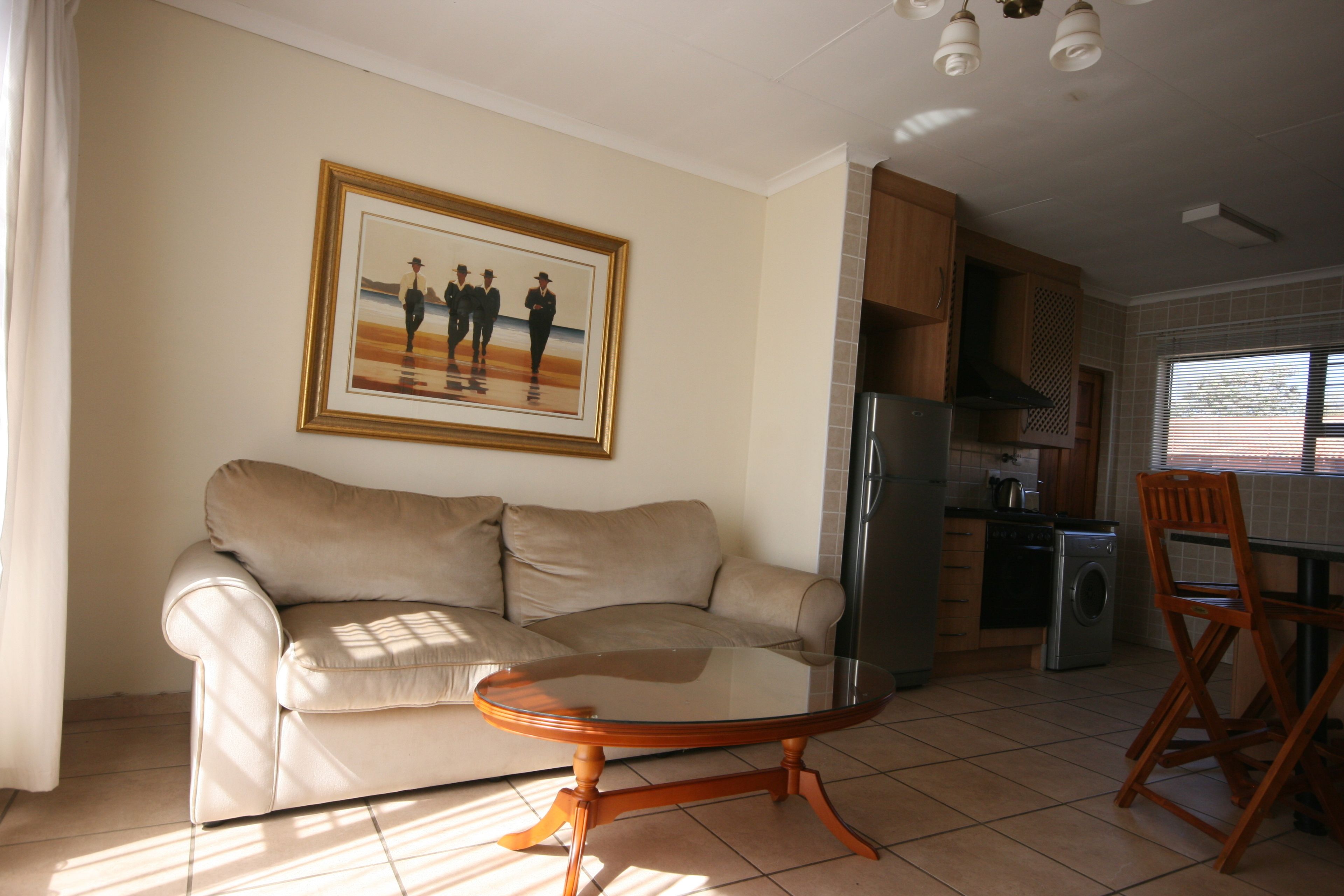 A living room with a beige sofa, a glass-top coffee table, a framed picture on the wall, and a kitchen area with cabinets and appliances.