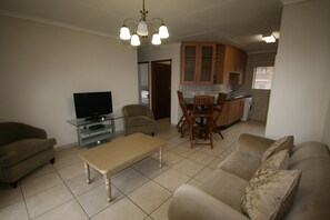 Una sala de estar con un sofá, una mesa de centro y un televisor en un mueble de vidrio. Hay un comedor con sillas y una cocina al fondo.
