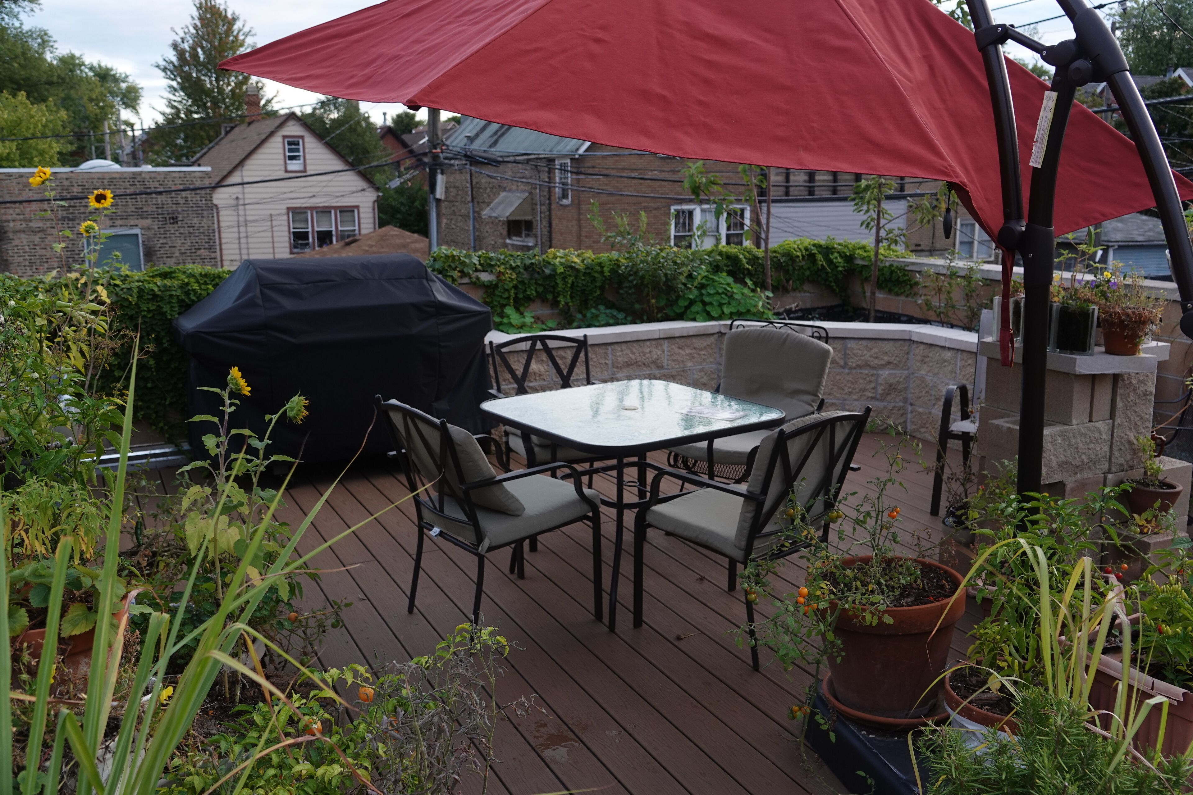 Een terras buiten met een tafel en stoelen, omringd door potplanten en een rode parasol.