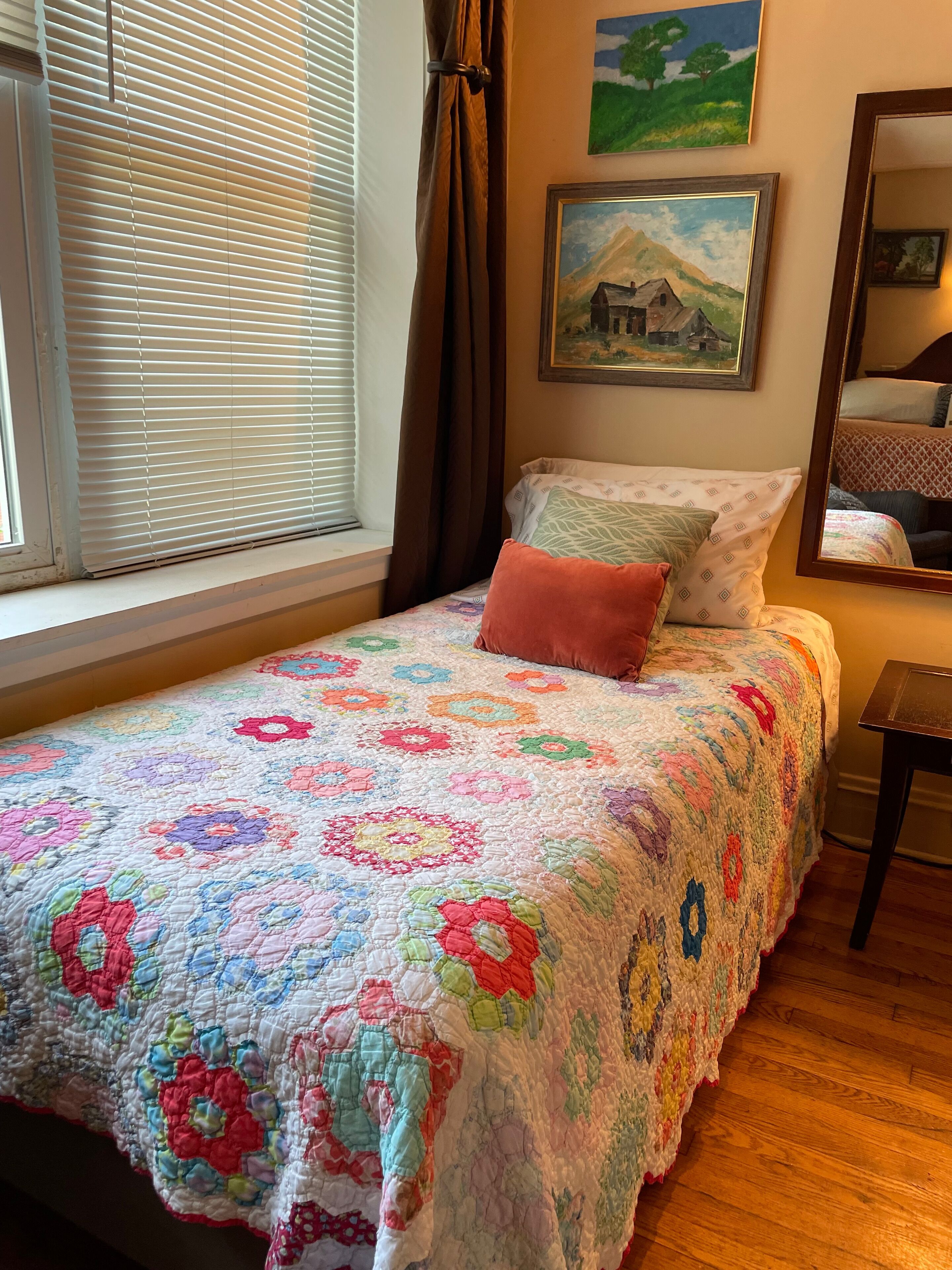 Un dormitorio con colcha floral, ventana con persianas, un cuadro y un espejo.