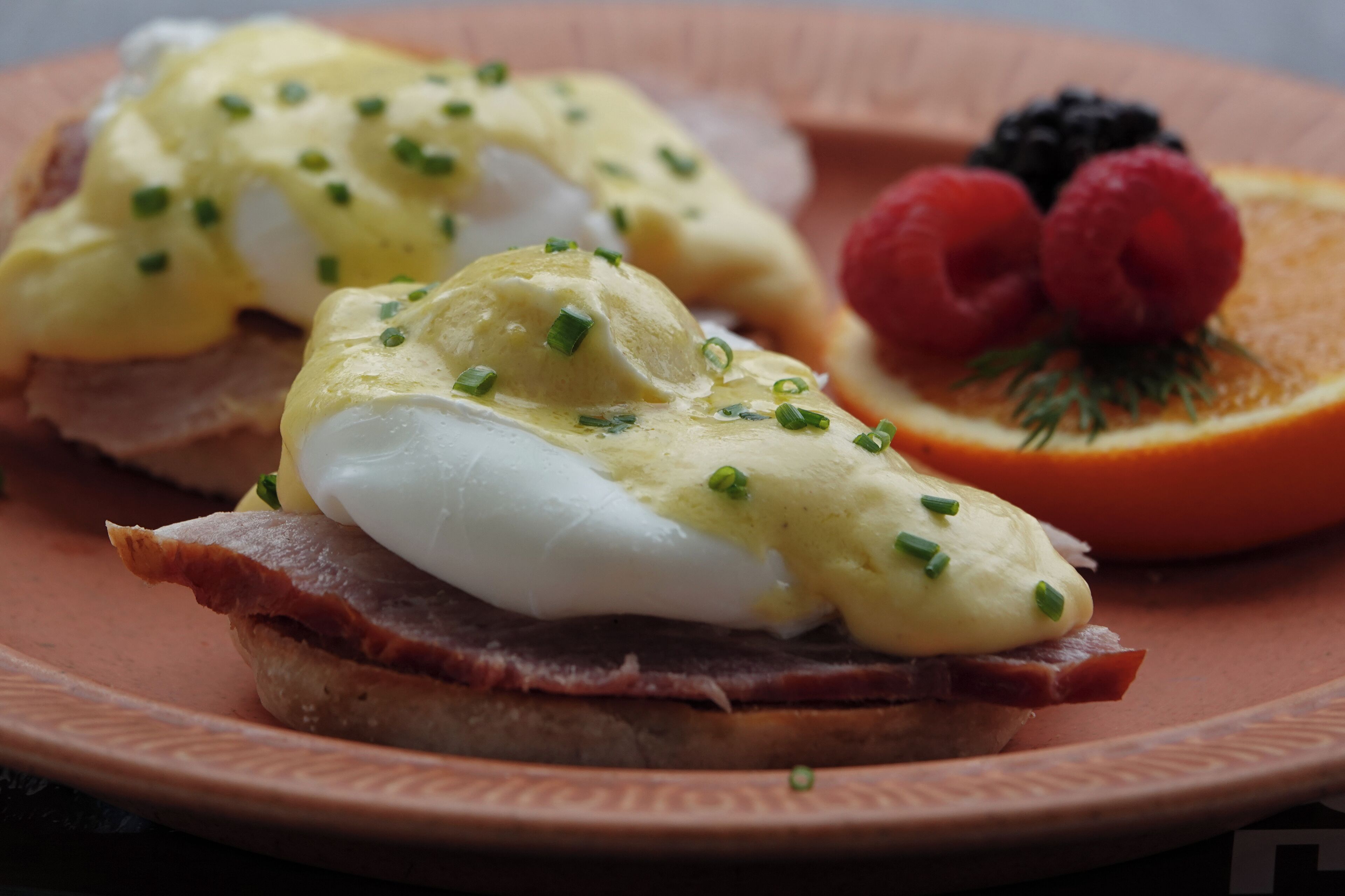 Un plato con huevos Benedictinos, jamón y fruta.