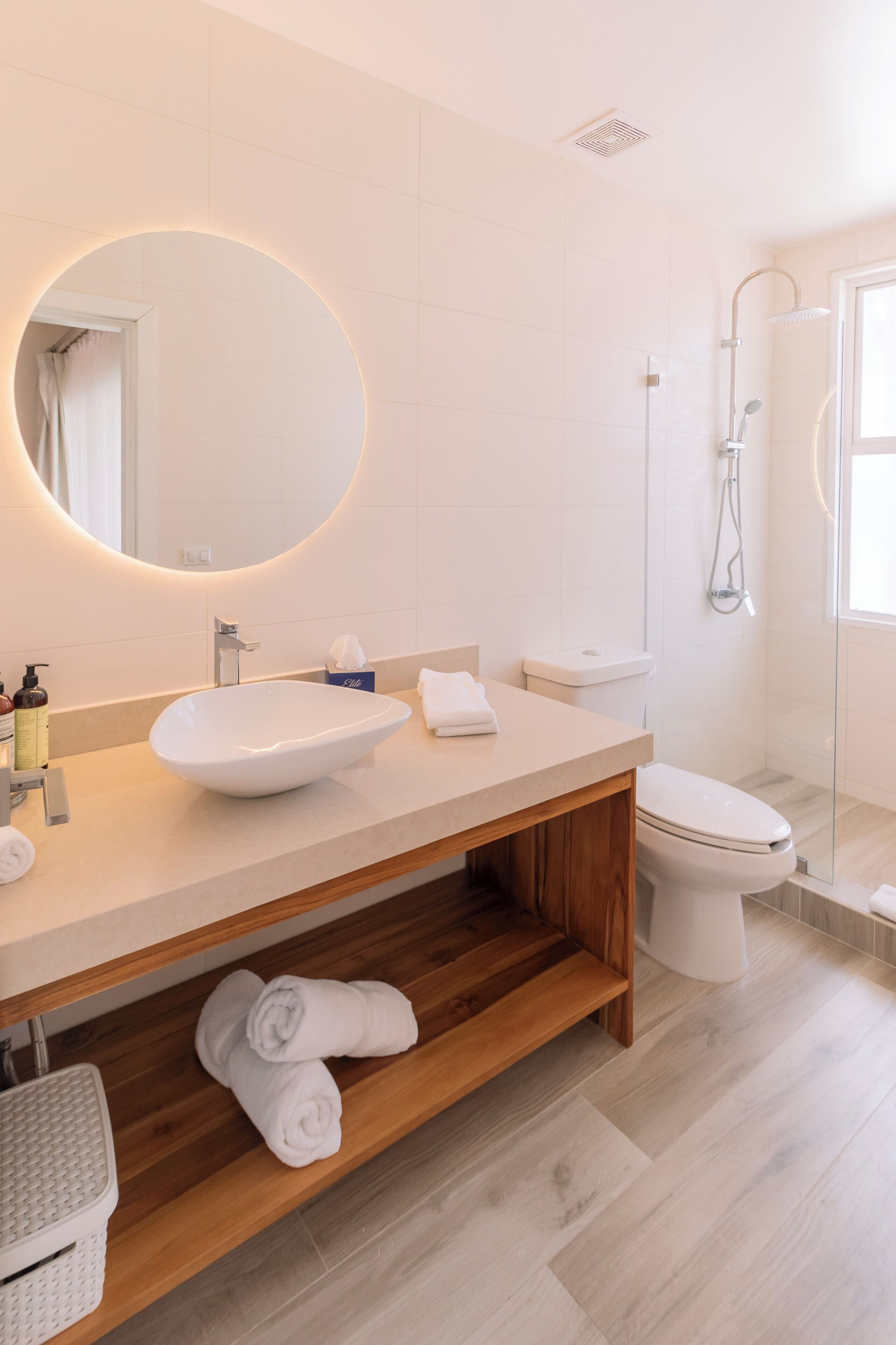 Un baño moderno con un espejo ovalado grande, un lavabo tipo recipiente, un inodoro y una ducha con mampara de vidrio.