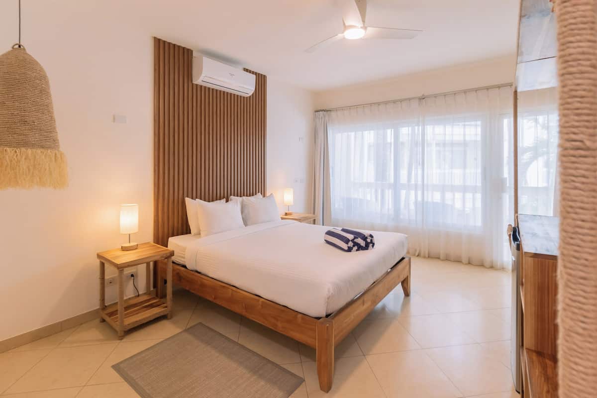 A neatly arranged bedroom with a wooden bed, a bedside table with a lamp, a ceiling fan, and a window with curtains.